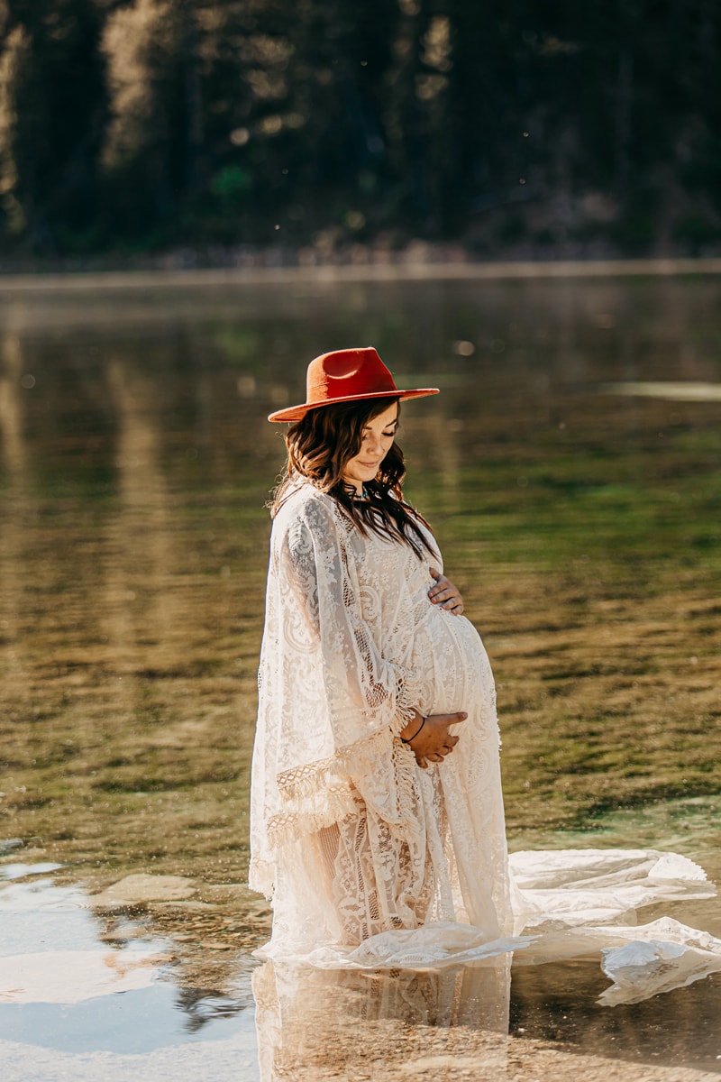 Maternity Photographer, a young mother-to-be wears a white gown and red hat stand in shallow lake waters