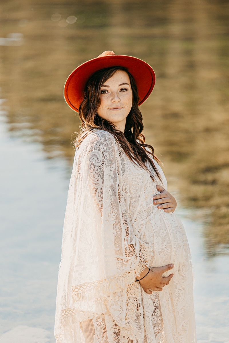 Maternity Photographer, a young expectant mother holds her belly and wears a white gown near the lake