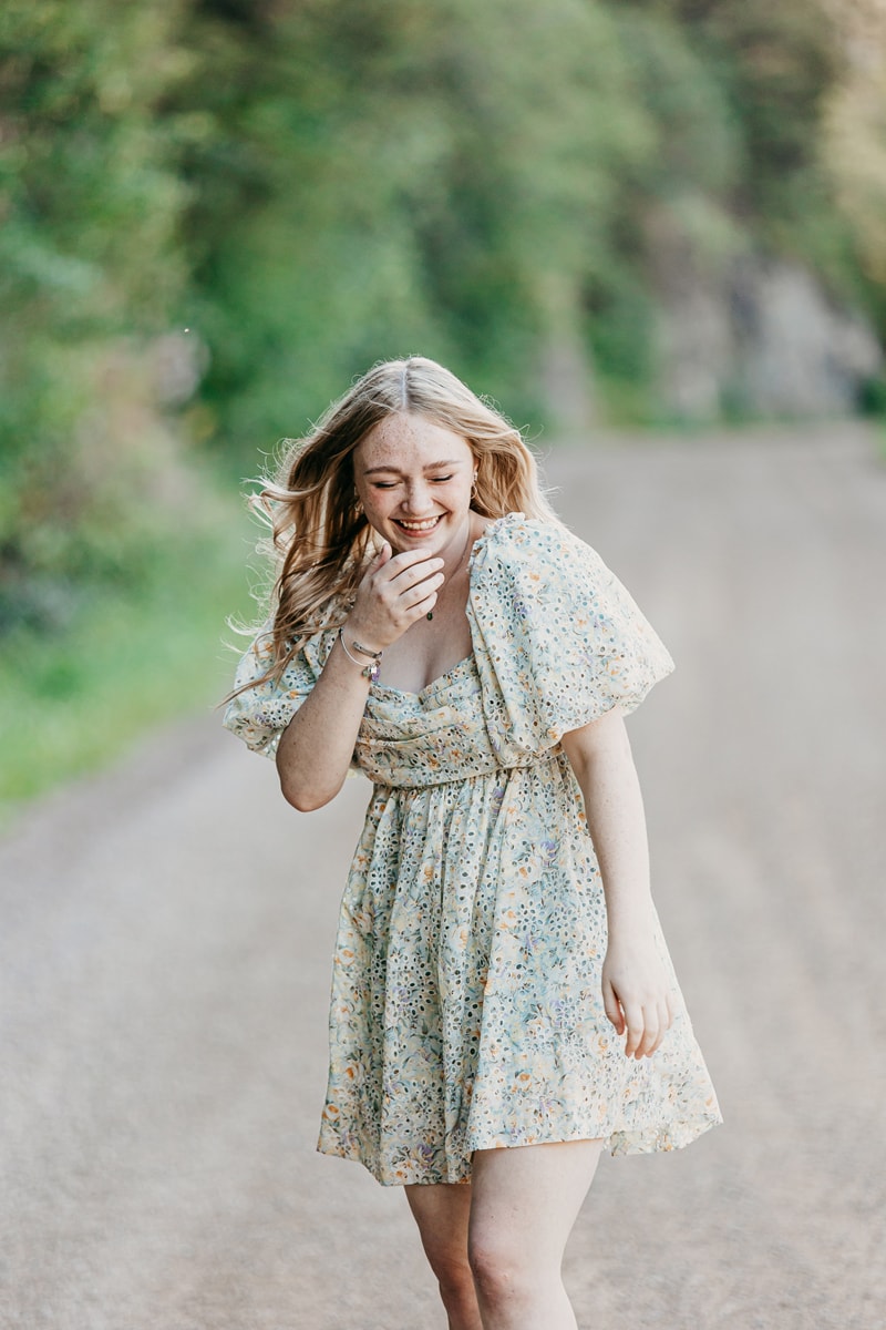 Senior Photographer, a young woman in a dress giggles as she walks down a country road
