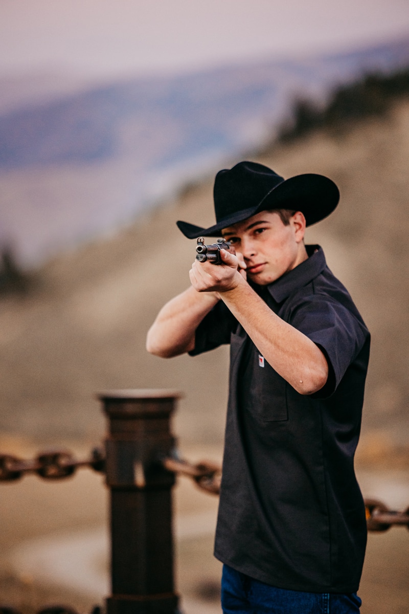 Senior Photographer, a young high school man holds a rifle and aims in dry meadows