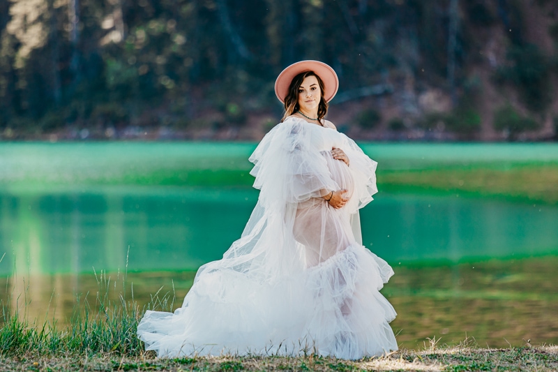 Maternity Photographer, a pregnant woman in a white gown stands before a lake