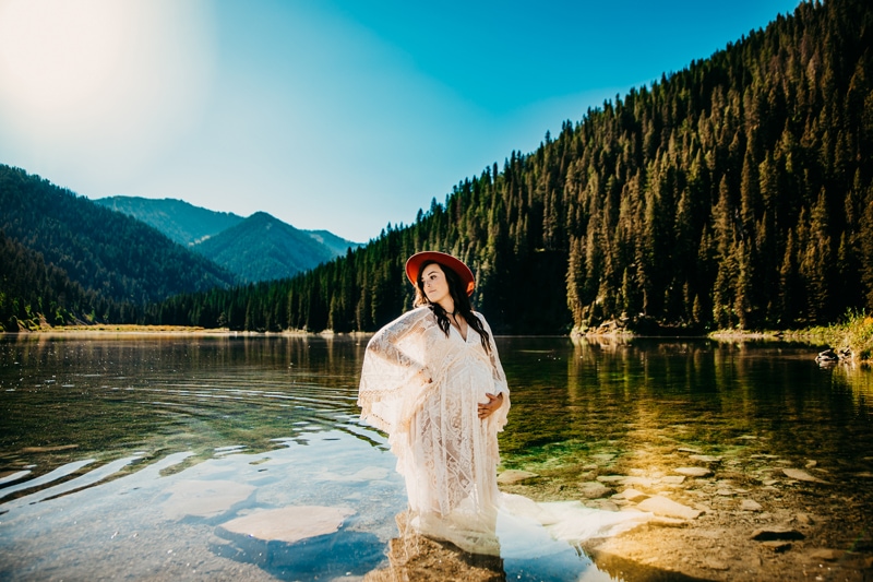 Maternity Photographer, a mother-to-be stands lake waters near mountain forests