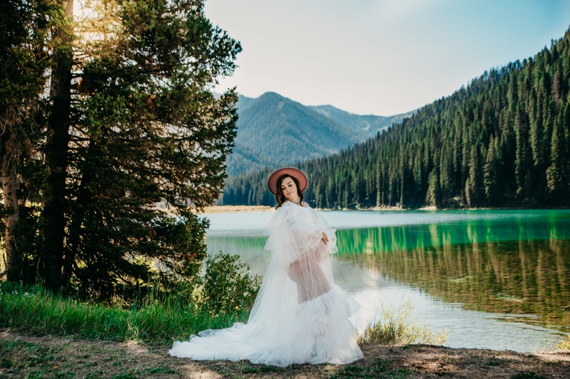 Maternity Photographer, a young mother-to-be stands near lake waters, she wears a hat and gown