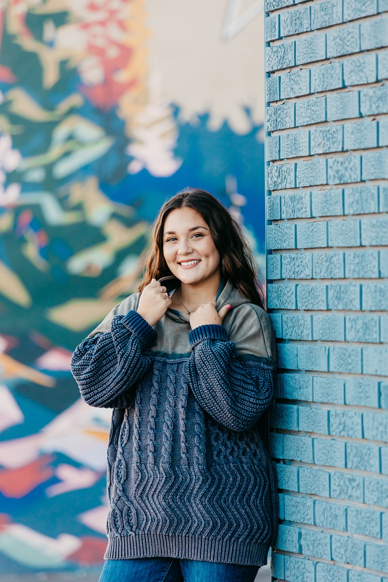 Senior Photographer, a young woman leans on a blue brick wall near a mural wearing a knit sweater and a smile