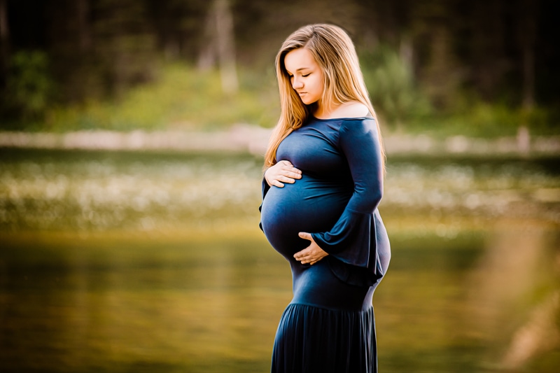 Maternity Photographer, a young mother holds her pregnant belly near a lake forest