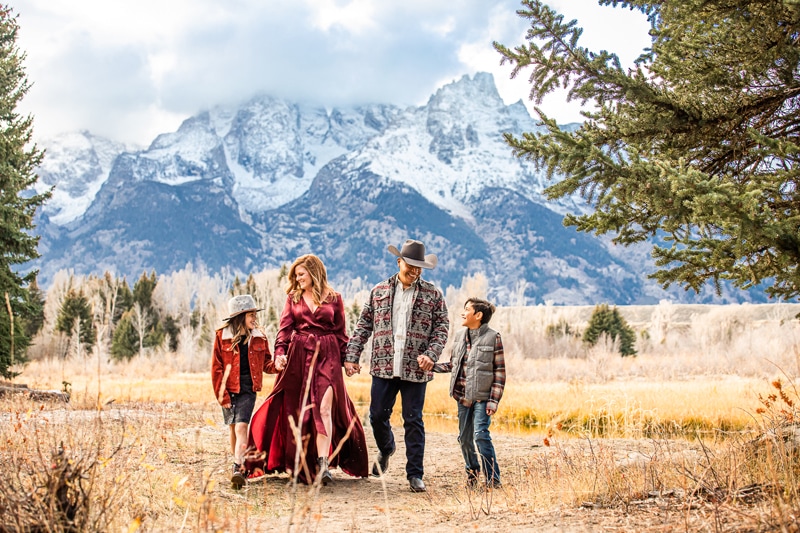 Family Photographer, a mother and father hold hands with their children in the meadows beneath snowy mountains