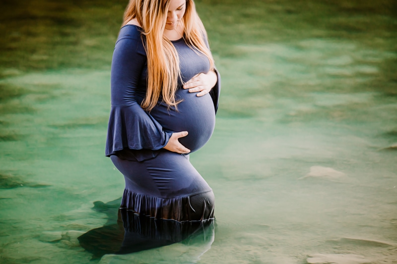 Maternity Photographer, an expectant mother holds her belly as she stands in shallow lake waters