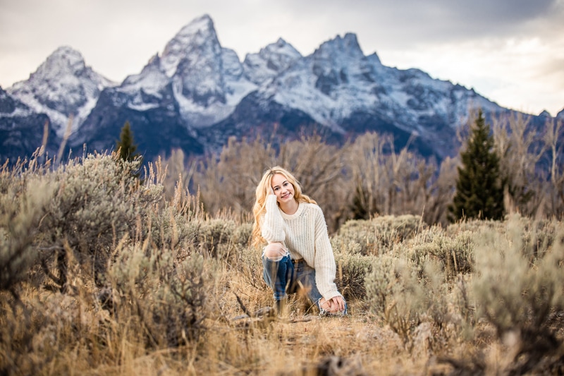 Senior Photographer, a high school woman kneels before snow topped mountains behind her