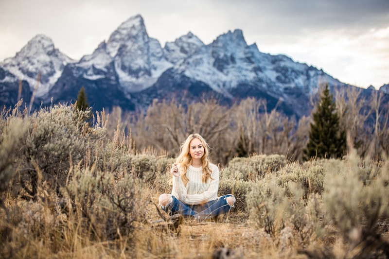 Senior Photographer, a high school woman sits in a dry chaparral near snowy mountain peaks
