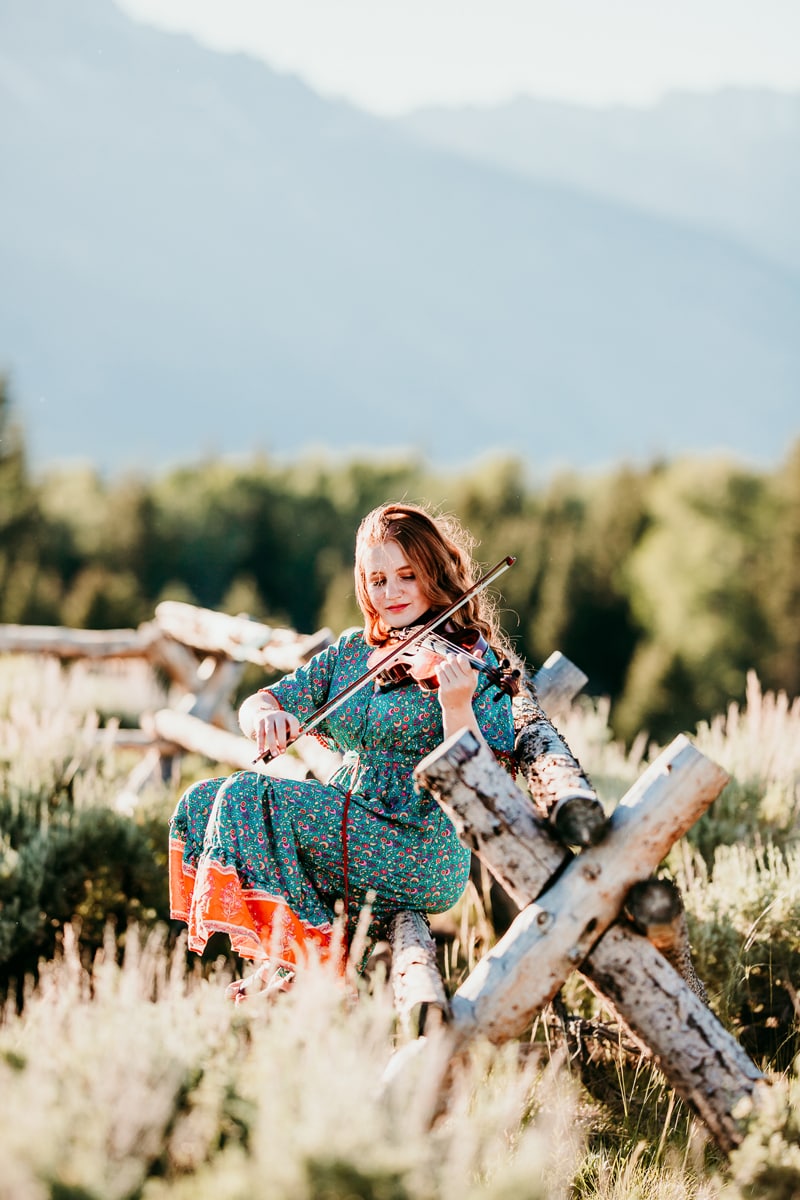 Senior Photographer, a high school aged woman sits in mountain meadows and plays the violin