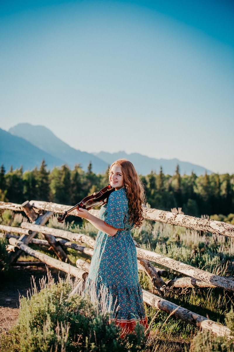 Senior Photographer, a young woman plays the violin and smiles as she stands next to a wooden fence near forest and mountains